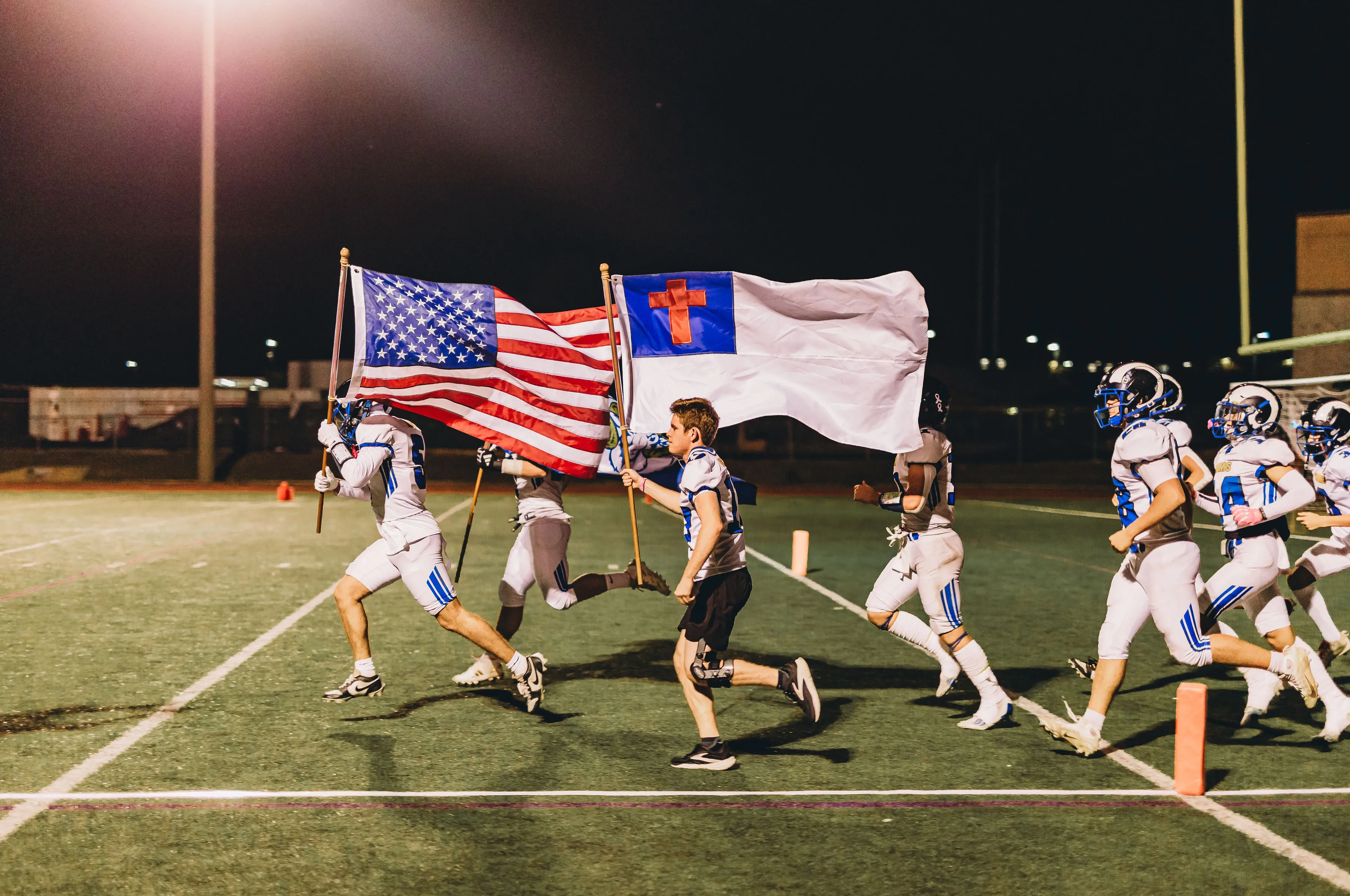 Rams football players taking the field carrying the American and Christian flags