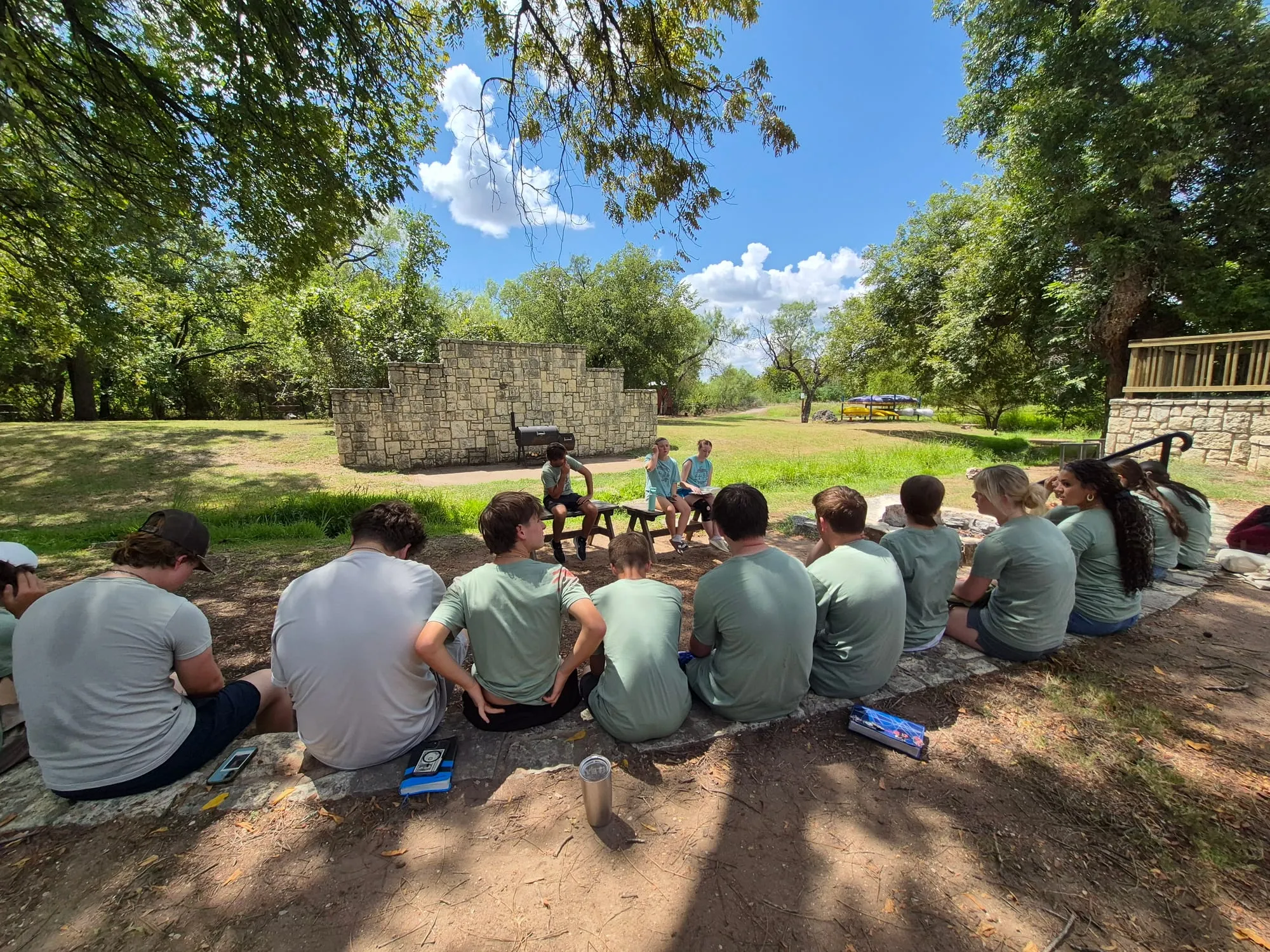 HCCS high school students sitting together outdoors at their retreat, listening to classmates speaking from a picnic table